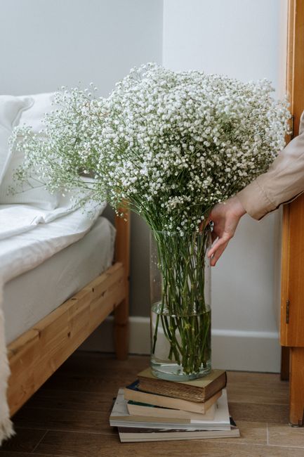 A large, elegant bouquet of fresh white Gypsophila, also known as baby's breath, arranged in a tall, clear glass vase filled with water. The bouquet is placed on top of a stack of books on the wooden