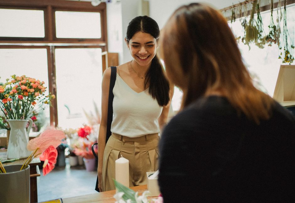 A young woman with long dark hair, wearing a sleeveless white top and beige high-waisted trousers, is smiling while standing in a flower shop, engaging in a floral arrangement or conversation with ano