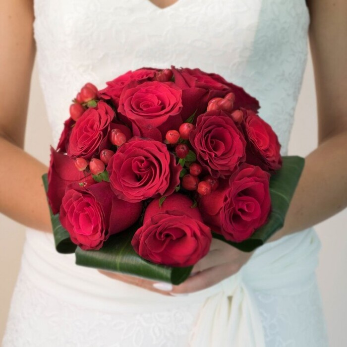 In this image, a bride is captured from the torso down, standing still against a soft, neutral background, her attention seemingly centred on the bouquet she holds carefully in both hands. The bridal bouquet is a striking, perfectly rounded cluster of deep crimson roses, so densely packed that their velvety blooms create a smooth, luxurious dome. Some of the roses still hold their petals close, bud-like and full of promise, while others are open and generous, displaying the intricate spiral at their heart where the petals overlap. Scattered throughout the roses are small bunches of bright red hypericum berries, each berry glistening slightly as if freshly polished, bringing a playful texture and a touch of festive brightness to the rich red palette. Encircling the base is a precisely arranged collar of broad, dark green leaves, folded back to frame the bouquet like a tailored cuff and to hide the stems within a clean, structured finish. The bride's hands, with neatly manicured nails in a soft, natural shade, support the bouquet from underneath, holding it close to the lace-covered bodice of her white gown. The dress itself features delicate floral lace detailing and a gentle, flowing skirt, a style common in bridal boutiques around Stanmore Broadway and The Broadway Circle. Soft, even lighting falls across the scene, accentuating the velvet texture of each petal and the glossy sheen of the foliage without casting harsh shadows. With the background blurred into creamy tones, the eye remains fixed on the vivid interplay of red and green. The overall effect is one of intense romance and timeless elegance, a classic red rose bouquet perfectly suited to a Stanmore wedding, whether exchanged in a quiet local chapel or a grand reception venue near Bentley Priory.