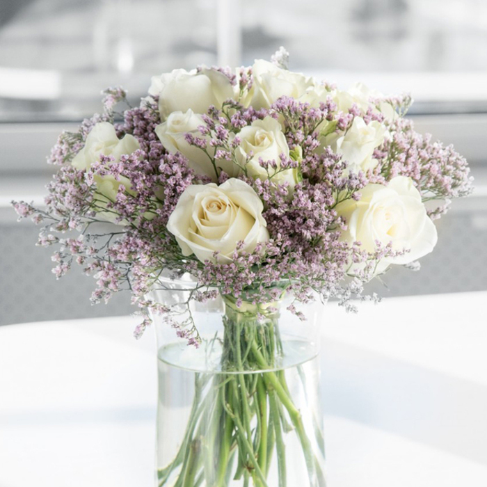 In this softly lit image, an elegant sympathy bouquet of white roses and lavender filler flowers is displayed in a tall clear glass vase, evoking the calm of a peaceful afternoon in Stanmore. The bouquet is composed mainly of velvety white roses, around a dozen in total, each one fully open and revealing its intricate spiral of petals. The inner petals are tinged with the faintest hint of warm ivory, gradually fading to a purer white at the edges, creating a subtle play of tone that adds depth and refinement. The blooms are clustered together to form a rounded, lush silhouette, yet each rose retains its own quiet presence, reflecting both purity and reverence. Surrounding and weaving between the roses are clusters of small, delicate filler flowers in soft lavender and pale lilac-pink shades, likely statice or limonium. Their many tiny, papery florets create a fine, lace-like texture that softens the overall look of the arrangement, giving it a cloud-like halo of gentle colour. The bouquet is anchored in a tall cylindrical glass vase filled with clear, fresh water, through which the straight, green stems are clearly visible, lined up neatly and extending downwards. This transparency highlights the freshness of the flowers and the care with which the arrangement has been made, reminiscent of the attention to detail offered by Flowers Stanmore for local condolence deliveries around Stanmore Hill and Old Church Lane. The vase sits on a smooth white surface that reflects some of the soft natural light bathing the scene. The background is intentionally out of focus, rendered in muted whites and greys that suggest a calm, modern interior-perhaps a quiet room where family or friends gather to remember a loved one. The overall atmosphere is soothing, dignified, and gently supportive, making this bouquet an ideal visual expression of sympathy and remembrance.