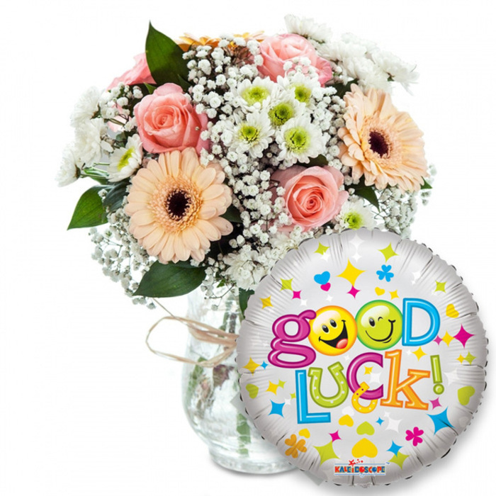 Against a simple white background, this thoughtful Good Luck gift set reads like a warm, visual note of encouragement from a neighbourhood florist. On the left and towards centre stage, a lush bouquet is artfully arranged in a clear glass vase tied with a piece of rustic raffia; the stems are visible through the glass, and the arrangement sits low enough to feel intimate. The palette is soft and optimistic: light-pink roses with layered, velvet petals, peach-toned Gerbera daisies whose dark brown centres ground the colour, and small white-and-lime green chrysanthemum-like florets that add lively points of brightness. Sprays of fine white gypsophila weave through the blooms like lace, while deep green foliage provides shadow and sculptural lines. In the foreground to the right, a round silver foil balloon overlaps the vase and repeats the upbeat message Good Luck! in a playful, multicoloured type. The two o's in Good are bright yellow smiley faces-one winking, one sticking its tongue out-bringing a wink of humour to the hopeful phrase; tiny stars, hearts and confetti in blue, pink, yellow and green decorate the balloon's metallic field. The overall feeling is encouraging and quietly festive, with the soft perfume of roses balanced by the crisp green note of chrysanthemums; it's an ideal condolence of optimism for farewells, graduations or interview send-offs around Stanmore Common and the Broadway.