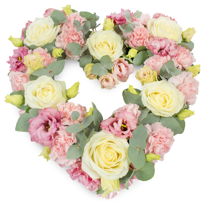 A soft, romantic heart-shaped wreath is shown against a crisp white background, its pastel flowers arranged with the thoughtful craftsmanship you might expect from Flowers Stanmore preparing tributes for local services. The heart is full and rounded, with a distinct inner curve and pointed base, yet no part of the frame is visible because every surface is cloaked in petals and foliage. Creamy ivory and light yellow roses are placed as anchor blooms throughout the design, their velvety petals spiralling outward from a tight centre in orderly layers, giving the arrangement an air of classic elegance. Surrounding and connecting these roses are generous clusters of light pink carnations, their numerous frilled petals creating depth and a soft, tactile feel that suggests comfort. Among the pastel carnation clouds, deeper magenta and mauve lisianthus blooms appear in thoughtful intervals, their gently flared petals and slightly darker hearts adding visual interest and a more emotive note, like cherished moments standing out among years of shared memories. Silvery-green eucalyptus leaves wind delicately through the floral mass, their rounded, satin-textured foliage introducing calm contrast and quietly tracing the heart's outer edge and inner dip. Tiny yellow-green buds peek out from the flowers, offering a touch of vitality and hinting that some blossoms are just beginning to unfurl. The overall palette of cream, pink, yellow, and muted green feels serene and soothing, perfectly suited to funeral or sympathy occasions in and around Stanmore, from quiet family gatherings to more formal services. The mood is one of affectionate remembrance and gentle farewell, carried entirely through the language of flowers. 