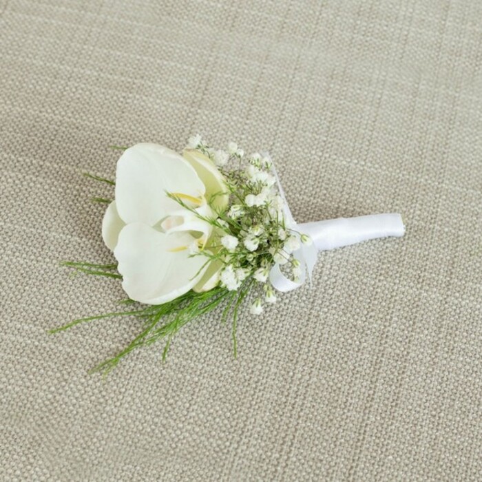 A meticulously crafted white orchid boutonniere lies on a neutral, light beige fabric with a clearly visible woven texture, similar to natural linen often used on tables in Stanmore's intimate wedding venues. The background's soft oatmeal tone and subtle weave create a calm, understated frame, allowing the floral details to stand out with quiet clarity. At the heart of the piece is a flawless Phalaenopsis orchid bloom, its petals smooth and gently arched, forming a serene, symmetrical face. The petals are a cool, creamy white, with a faint, almost translucent quality along the edges, while the inner throat reveals delicate hints of pale yellow and soft cream, adding warmth and depth to the centre of the flower. Encircling the orchid, dainty clusters of white baby's breath form a soft cloud, the tiny round blossoms dotted along fine stems. Their texture is light and frothy, creating a gentle contrast to the broad, sleek petals of the orchid and giving the boutonniere a graceful, airy silhouette. Fine strands of slender green foliage peek out from beneath and between the flowers, adding a fresh, botanical note; their feathery structure implies a gentle movement, as though just gathered from a garden near Belmont Circle or along the leafy roads leading through Stanmore. The stems of the orchid, gypsophila, and greenery are neatly aligned and tightly wrapped in a matte white satin ribbon. The ribbon is smooth and immaculate, wound evenly around the base and finished with a discreet, tidy bow that sits close to the stems, demonstrating the florist's careful hand. Soft, diffuse lighting washes over the arrangement, highlighting the contrast between the crisp whites and vibrant greens without casting harsh shadows. The overall effect is one of classic elegance and modern minimalism, an ideal boutonniere for a groom or groomsman at a Stanmore ceremony, capturing purity, fresh beginnings, and the subtle sophistication that local wedding flowers can bring to a perfectly tailored suit.