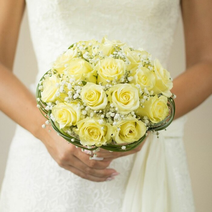 A carefully composed close-up from the vibrant heart of Stanmore captures the poetic beauty of a bridal bouquet held close, its colours radiant against the gown's detailed white fabric. Exquisite pale yellow roses, each perfectly blossomed, rise in harmonious domes, their soft petals glowing as if kissed by the afternoon sun near Canons Park. Miniature galaxies of white baby's breath weave between the blooms, lending an ethereal delicacy and gentle movement to the bouquet, reminiscent of the gentle breezes wafting through Stanmore's leafy avenues. At the base, swathes of verdant green leaves and fine decorative wire curl in intricate, graceful loops, defining the structure and elegance expected from a skilled Stanmore florist. The hands gently supporting the arrangement, with softly manicured nails and a wedding ring glimmering modestly, radiate both care and pride. The softened, neutral backdrop draws all focus to the connecting beauty of the bride and her bouquet, a scene overflowing with the promise and joy of a wedding celebrated here in Stanmore. This image perfectly reflects the enduring romance and refined artistry that couples seek for their most important day, bringing together local tradition and timeless style in a single, unforgettable floral centerpiece.