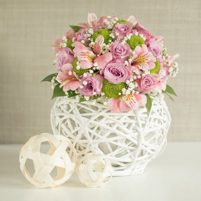 This refined floral arrangement, dazzling in its sophistication, channels the character of Stanmore's elegant homes and garden weddings. Centered on a bright white surface, the bouquet overflows with pastel pink and lavender roses-lush and fully open-framed by long-lasting pink alstroemeria whose delicate trumpet blooms are streaked with gentle yellow and blush markings. Compact, lime green button chrysanthemums scatter bursts of lively colour, adding both structure and contemporary vibrancy. Airy white baby's breath delicately weaves through the composition, softening each transition and adding a layer of ethereal charm. Cradling the arrangement is an eye-catching white vase, its open-weave design sculpted from intricate, slender strips-a crafted look frequently found in Stanmore's design boutiques and artisan fairs. Its broad, rounded base exudes substance and style, forming a perfect stage for this floral celebration. Adjacent, two woven decorative spheres in soft, natural tones complement the vase's intricate structure, grounding the visual composition in tactile artistry. The subdued beige background, textured like fine woven linen, ensures the flowers and their vessel remain the star. Illuminated by gentle daylight, this centerpiece radiates the warmth and joyous refinement that distinguishes Stanmore's social gatherings, and is effortlessly suited for commemorating weddings, anniversaries, or special family occasions.