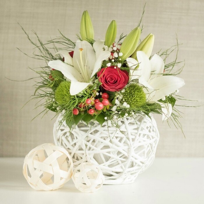 This image presents a refined floral arrangement that would make a striking wedding table centerpiece in Stanmore, marrying classic romance with contemporary style. The flowers are gathered in a rounded white vase whose open-weave structure resembles gently intertwined branches or wicker, lending an airy, almost sculptural quality to the base. Resting on a pristine white surface, the vase appears to glow subtly, echoing the light tones of the background wall, which is a soft, neutral beige with a delicate, barely noticeable texture. Rising from the vase is a generous display of white lilies whose elongated petals curve gracefully outward, some fully open and others still unfurling. The open lilies reveal pale speckles and confident yellow stamens, conjuring a sense of purity and brightness well suited to wedding celebrations around Stanmore, whether in a local hall or a marquee set up near the green spaces. Interlaced with these lilies are several red roses, their velvety petals tightly arranged into spirals that pull the eye inward; a central rose, placed near the front of the design, provides the main focal point and an unmistakable symbol of love. Between these larger blooms, bright green pom-pom flowers-likely green chrysanthemums or dianthus-introduce playful spheres of colour and touchable texture, standing out vividly against the white petals and rich crimson tones. Delicate sprays of white baby's breath thread through the arrangement, softening edges and adding an ethereal, cloud-like layer. Small red berries punctuate the greenery with warm, festive accents, making this centerpiece well suited to winter and holiday weddings. A mix of fine, wispy foliage and more robust dark green leaves form a lush, layered backdrop, draping slightly over the rim of the vase and suggesting movement. To the left of the arrangement, two decorative spheres made from pale woven material rest on the tabletop; they echo the vase's open lattice pattern, completing a cohesive and stylish scene that feels as though it has been thoughtfully arranged by an expert Stanmore florist who understands how to make a wedding table feel both special and welcoming.
