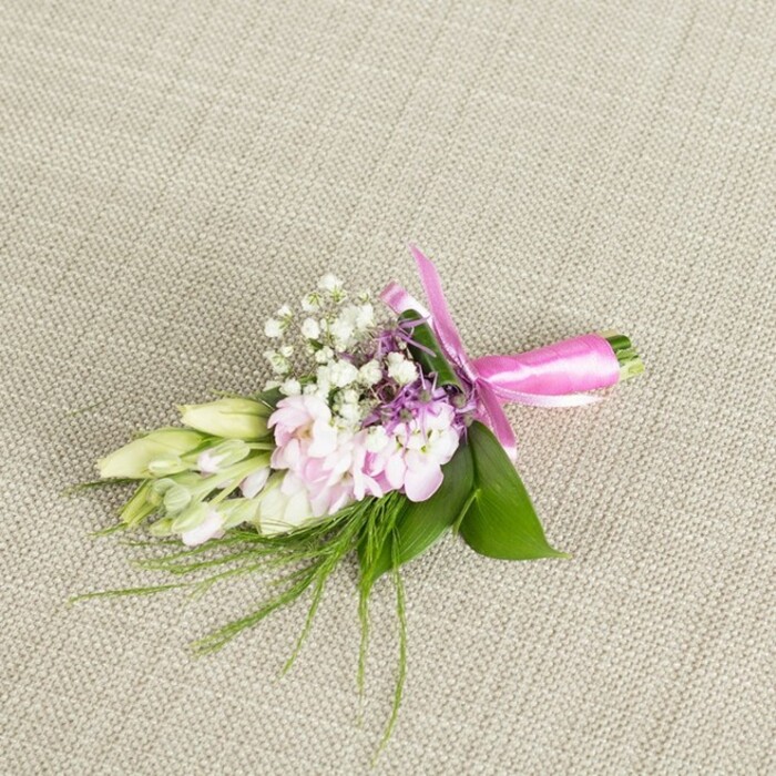 A meticulously arranged floral boutonniere or petite corsage lies centered on a neutral, light greige linen backdrop, the fine weave of the fabric adding a subtle, tactile context that suggests understated luxury often favoured by couples marrying around Stanmore and the wider HA7 area. The composition is anchored by a cluster of soft, light pink blossoms, their petals gently ruffled and layered, reminiscent of miniature stocks or delicate garden blooms gathered at their peak. Each flower reveals a faint gradient of colour, with slightly deeper notes nestled in the heart and paler tones at the edges, giving the arrangement a soft-focus, romantic quality. Interlaced among these focal flowers are airy sprays of white baby's breath, their tiny round florets forming a delicate mist that lightens and expands the visual volume, like a fine veil across the blooms. Small accents of lavender and light purple florets emerge between the petals, lending a hint of depth and bringing a subtle, contemporary twist to the traditional pink-and-white palette. To one side, a line of slender, tapered buds in creamy white and pale green extends gracefully outward, suggestive of freesias or similar buds that have not yet opened, symbolising anticipation and new beginnings. A foundation of rich, vibrant green leaves cradles the flowers, their broad surfaces providing contrast, while fine, wispy, grass-like greenery and ferny sprigs dance outwards from the base, adding a slight wildness, as though freshly cut from a garden near Whitchurch Lane. The stems are carefully bound together and finished with a hot pink, fuchsia satin ribbon, tied into a refined, symmetrical bow whose glossy surface catches the light and introduces a playful, celebratory note. The overall effect is of a fresh, handcrafted accessory, perfect for wedding buttonholes, prom outfits, or special family celebrations in and around Stanmore, where attention to delicate detail matters.