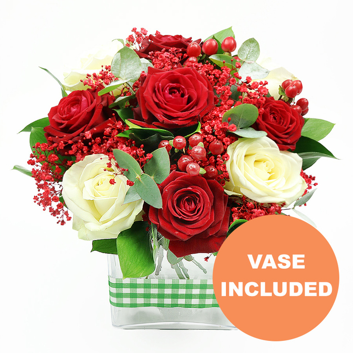 A richly coloured floral arrangement commands attention against a clean white background, reminiscent of a carefully styled centrepiece in a Stanmore living room on a special evening. At its heart, deep red roses, full and velvety, open wide to reveal layers of rich, crimson petals, exuding warmth and passion. Interspersed among them are creamy white roses, their soft, ivory tones bringing light and gentle contrast, lending the bouquet an air of timeless romance. The spaces between these classic blooms are filled with fine sprays of tiny, bright red filler flowers, similar in form to gypsophila; they form airy, delicate clusters that soften the edges of the roses and give the composition a slightly misty, celebratory feel. Nestled between petals, rounded red hypericum berries catch the eye with their glossy surfaces, adding playful dots of colour and textural interest. A mixture of fresh, leafy greenery surrounds the roses: elegant eucalyptus leaves with their subtle silvery-green cast, along with deeper green foliage that fills and frames the bouquet, enhancing its volume and giving it a lush, garden-like presence, much like the foliage you might see walking near Bentley Priory Nature Reserve. The entire display is gathered neatly into a clear rectangular glass vase, where the stems can be seen running straight down into the water, reinforcing the sense of clean lines and modern style. Around the middle of the vase, a green and white gingham ribbon is wrapped in a tidy band, its checked design adding a rustic, kitchen-table charm to the otherwise polished arrangement. In the lower right corner of the image, an orange circular badge with bold white capital letters announces VASE INCLUDED, clearly indicating that the bouquet arrives as a complete, ready-to-place gift, ideal for same-day delivery throughout Stanmore for occasions like anniversaries, festive dinners, or simply to bring a touch of elegance and warmth into someone's home.