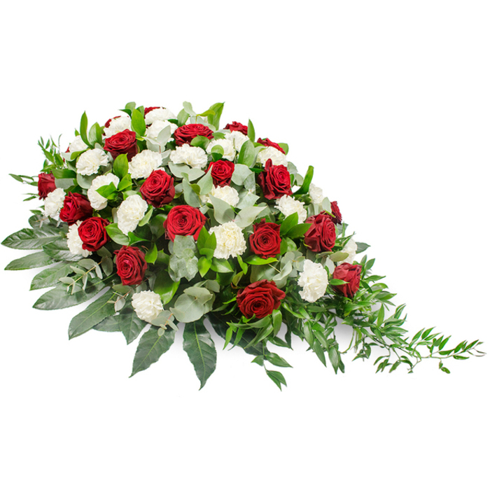 An elegant funeral flower spray, designed in classic tones of deep red and white, is photographed against a pure white backdrop that emphasises its shape and detail. The arrangement has a horizontal, slightly asymmetrical form: the left side appears more rounded and lush, while the right side narrows gracefully into a subtle, trailing point, creating a sense of quiet movement. Deep crimson red roses are nestled throughout, their velvety petals densely layered and rich with colour, some blooms set a little higher to give gentle variation in height. Between them, an abundance of white carnations fills the spaces, the ruffled texture of their petals creating a soft, cloud-like effect that contrasts beautifully with the smooth roses. Together, the red and white flowers convey love, reverence, and a wish for peace, making this spray especially suited to funerals and memorial services. The flowers are supported by a generous base of greenery: broad, dark green leaves provide structure, shaping the outline of the piece, while pale, silvery-green eucalyptus leaves weave in and out, catching the light and introducing a delicate, aromatic freshness. On the right-hand end, slender, trailing stems of lighter green foliage, likely ruscus or similar, extend outward and slightly downward, giving the whole design an easy, flowing finish. The spray feels thoughtfully composed, like a tribute prepared by Flowers Stanmore for a local family saying farewell at a service near Stanmore station or in a nearby place of worship. It is the kind of memorial arrangement chosen to rest on a casket or remembrance table, expressing condolences with grace, tradition, and a personal, local touch.
