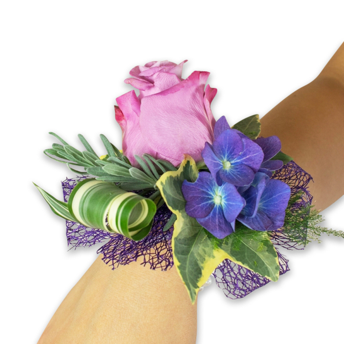 Laid diagonally across a light-toned forearm and photographed on white, this wrist corsage is a study in refined contrasts and careful craft-one I would happily prepare at Flowers Stanmore for a wedding or special evening in the locality. The central bloom is a generously sized, partially open pink rose whose petals graduate from a deep fuchsia-rim to a softer blush toward the heart, each layer plush and slightly luminous. Surrounding it are three prominent clusters of deep indigo-purple hydrangea, their tiny blossoms forming a textured mass with subtly blue-tinted centres that seem to glow against the rose's warmth. Silver-green sprigs-evoking eucalyptus or dried lavender-create a soft, feathery backdrop, while glossy variegated ivy with dark green centres edged in creamy yellow adds a timeless, garden-like frame. A bold, broad leaf with white and green stripes is artfully curled into a spiral for a modern sculptural line, balancing the arrangement's softness. Beneath, a layer of open-weave dark purple mesh peeks through, increasing depth and amplifying the violet tones. The lighting is soft and natural, bringing out the tactile qualities: the rose's velvet, the hydrangea's papery clusters, the ivy's sheen, and the mesh's delicate shadow play. Whether worn for prom, a bride's party, or a celebratory reception near Bentley Priory or Stanmore Broadway, this corsage reads as elegant and composed-freshly made, locally considerate, and designed to feel both contemporary and timeless.
