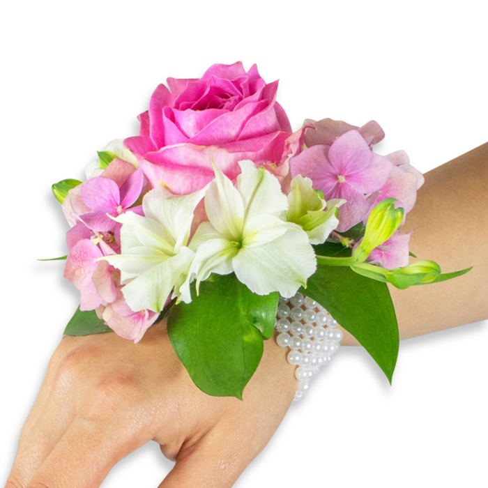 An elegantly composed wrist corsage presented on a light-to-medium skin toned left hand, photographed against a stark white background that sets every hue and texture in crisp relief. The focal point is a large, fully bloomed hot fuchsia-pink rose, its dense, velvety layers arranged with the precision of a practiced hand. Nestled around the rose are clusters of soft, blush pink hydrangea florets that give the design a generous, tactile fullness, while pristine white alstroemeria (Peruvian lilies)-their centers subtly streaked with pale green-offer bright, linear accents that lift the palette. A cluster of pale green alstroemeria buds and several smaller, nearly closed pink buds sit to the right, suggesting the living quality of the arrangement and the patience of bloom. Broad, rich emerald leaves form a fresh, structured cradle beneath the flowers, providing contrast in both colour and sheen. The entire arrangement is secured to the wrist with a refined multi-strand bracelet of small white pearls that wrap snugly around the wrist and trail partway down the back of the hand; the pearls' soft luminescence complements the flowers' highlights and speaks to careful construction-wired stems discreetly anchored, floral tape and padding kept invisible so the accessory remains comfortable to wear. Crafted locally by Flowers Stanmore on Stanmore Broadway, this corsage reflects neighbourhood sensibility and floristry skill, perfect for weddings, proms, or commemorative moments across Stanmore and nearby Harrow.