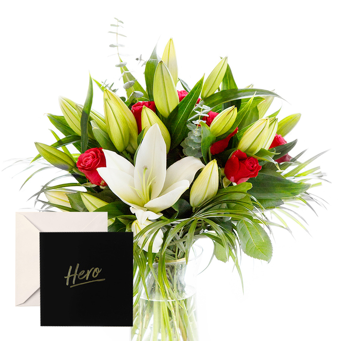 Like a modern gallery piece, this arrangement blends bold with serene. White lilies drift at the heart of the bouquet, luminous and airy, their satin petals contrasting splendidly with the tactile greenery that encircles them. Rich ruby roses punctuate the design, each bloom a symbol of love and respect as they rise among the stems. Verdant leaves provide a cooling, almost forest-like backdrop, while unopened lily buds suggest anticipation and new beginnings. In soft Stanmore daylight, the bouquet feels alive, almost breathing, and ready to lift the mood of any room. Each stem is placed with careful rhythm, creating depth and movement that guides the eye from the clear glass vase up to the crown of blooms. The fragrance sits lightly, a whisper of spring amid the quiet energy of a Stanmore home. Hints of eucalyptus and wispy grasses add a subtle herbal freshness, giving the arrangement motion without distraction. This piece is designed with gratitude and reverence, a vibrant tribute to someone cherished and extraordinary, whether marking a milestone, a thank you, or simply celebrating a personal hero. The Hero card anchors the meaning, and same-day delivery across Stanmore, Canons Park, and the local streets brings this refined bouquet to a doorstep near The Broadway.