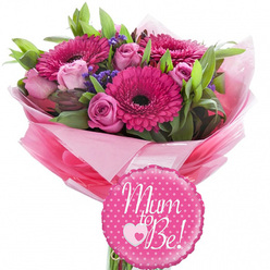 Against a pure white backdrop, this maternity-themed bouquet glows with shades of pink and fresh green, like a little celebration ready to be carried through the quiet streets around Stanmore Hill. The eye is immediately drawn to the vibrant fuchsia gerbera daisies, each one a confident burst of colour with a deep, inky centre and neatly layered petals that create a sense of joyful movement. Woven between them are soft pink roses, some tightly wrapped like delicate secrets, others gently opening to reveal velvety inner spirals, hinting at the unfolding journey of motherhood. Delicate purple filler flowers, likely statice or limonium, add a subtle haze of lilac, softening the overall look and giving the arrangement an airy, almost cloud-like texture. Lush greenery frames and supports the pink blooms, with slender leaves and broader, glossy foliage creating depth and a natural, garden-fresh feel. The stems are cocooned in layers of pastel and brighter pink tissue paper, with a sheer cellophane outer layer that catches reflections, suggesting careful, professional wrapping by a local Stanmore florist. At the base sits a round, scalloped-edge foil balloon in bright fuchsia, dotted with crisp white polka dots. Across the centre, in graceful white script, the phrase "Mum to Be!" is written, accented by a sweet white heart enclosing a smaller pink heart, echoing the bond between mother and baby. The overall mood is one of warm anticipation and gentle excitement, making this arrangement ideal for a baby shower, gender reveal, or to send congratulations to an expectant mum anywhere in Stanmore.