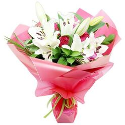 A luxurious floral arrangement of white lilies and deep red roses is shown against a bright white backdrop, every petal, leaf, and fold of wrapping clearly defined. The lilies, with their large, open trumpets and gently recurved petals, take centre stage, their pure white surfaces acting like soft reflectors of light. In the middle of each bloom, dark, chocolate-brown anthers sit atop slender filaments, their colour echoing the richness of the red roses below. Several lily buds, tall and slender with green outer casings, rise from the bouquet, promising continued blooms once the flowers are delivered to a home in Stanmore. Interwoven with the lilies, the red roses provide a velvety contrast, some buds tightly closed, others beginning to loosen into layered spirals that convey romance and devotion. Around and behind the blooms, abundant greenery in deep and mid-green tones fills the spaces, with broad, glossy leaves catching subtle highlights and fine, grass-like stems adding vertical lines and texture, reminiscent of the gentle sway of trees around Bentley Priory Nature Reserve. The whole composition is wrapped generously in shiny, bright pink cellophane that appears slightly translucent, its crinkled, ruched folds forming a flared collar around the flowers. At the point where the wrap narrows to hold the stems, a narrow lime green satin ribbon is tied, its crisp bow adding a playful yet refined touch. This bouquet feels both classic and contemporary, ideal for celebrating milestones, saying thank you, or sending love within Stanmore, and reflects the skill and attention to detail you would expect from a dedicated neighbourhood florist.