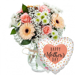 This photograph presents a tender Mother's Day gift set, thoughtfully composed and shown against a pristine white backdrop that echoes the calm, open feel of Stanmore's residential streets. Dominating the left side of the frame, a lush mixed bouquet rises from a clear glass vase, the stems visible through the water like a neat bundle gathered by an experienced florist from a studio near Stanmore Broadway. The vase is finished with a simple loop of rustic twine at its neck, adding a quiet, handcrafted note that suggests time and care have gone into this gift. The bouquet itself is rounded and bountiful, packed with soft, romantic colours. Fully opened peach and blush-pink roses showcase spirals of velvety petals, each bloom inviting you to picture the soft touch of the flower and the comfort of giving it to someone dear. Around them, bright peach gerberas with deep, dark centres bring in a more playful element, their wide, symmetrical faces reminiscent of the cheerful energy of a family gathering. Between the larger blooms, a light mist of white baby's breath softens the lines of the arrangement, creating a dreamy, cloud-like effect that makes the bouquet feel light and airy. Small white chrysanthemums with pale green hearts nestle in the gaps, adding texture and giving the arrangement resilience and shape, much like the layered greenery you might find on a walk through Bentley Priory Nature Reserve. Deep, glossy foliage surrounds and supports the flowers, enriching the palette and suggesting freshly conditioned stems. In the foreground to the right, a large heart-shaped foil balloon adds unmistakable celebration. Its white surface is sprinkled with metallic gold and silver dots, scattered like confetti tossed into the air. At the centre, a warm peach heart frames the words HAPPY Mother's DAY in bold black type, the word Mother's written in flowing, calligraphic script that brings intimacy to the message. A small white card tucked behind the vase and balloon hints at a personal note. Together, the bouquet, balloon, and card create a complete visual story of appreciation, ideal for a Mother's Day delivery to a home anywhere in Stanmore.