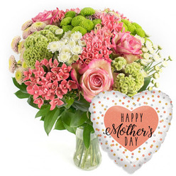 A bright, contemporary Mother's Day flower arrangement is the focus of this image, carefully photographed against a pristine white backdrop that is typical of online florist displays for areas such as Stanmore. In the centre sits a clear glass vase, its clean lines and visible stems giving a sense of freshness and simplicity. Emerging from the vase is a domed bouquet, dense with blooms and foliage. Large pink roses take pride of place, their petals unfolding in spirals and edged with a slightly darker blush that adds depth and tenderness. Nestled between them are small clusters of coral-pink blooms, much like kalanchoe or similar multi-flowered stems, bringing a lively, almost sparkling texture. Vibrant lime-green button chrysanthemums and velvety, frilled chartreuse flowers add an unexpected modern pop, their cool green tones standing out against the warmer pinks and corals. Sprinkled throughout are delicate white spray chrysanthemums and tiny daisy-shaped blossoms, which brighten the arrangement and give it a soft, uplifting feel, reminiscent of spring walks around Stanmore Country Park. Dark, glossy greenery provides structure and a rich base, ensuring the bouquet looks full and balanced from every angle. To the bottom right of the flowers hovers a heart-shaped foil balloon, its surface shining softly. The balloon's white background is patterned with irregular metallic dots in shimmering gold, silver, and peach. At its centre, a peach heart carries the words "HAPPY Mother's DAY" in black type, with a mix of bold and cursive styles that echo both celebration and affection. The overall composition feels warm, thoughtful, and festive, ideal for representing a complete Mother's Day gift of flowers and balloon delivered to homes across Stanmore.