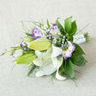 On a neutral, light beige fabric that resembles a well-loved linen tablecloth, a horizontal bouquet rests as if waiting to be carried down the aisle at a Stanmore ceremony. The composition centers on magnificent white Oriental lilies, their broad petals opening in graceful curves, revealing golden-orange stamens dusted with dark pollen and fine speckles that trail along the cream interiors. Some lilies remain tightly wrapped in lush green buds, adding both height and a promise of future blooms. Between these star-like flowers, lisianthus blossoms unfurl in white and purple, each petal delicately ruffled, transitioning from soft ivory at the throat to vibrant lilac and violet at the edges. Several buds hover at different stages of opening, bringing a sense of life and gentle movement to the design. Tiny clusters of baby's breath sit like soft clouds around the main blooms, their miniature white flowers adding a tender, romantic haze. Enveloping the arrangement is a wealth of greenery: broad, glossy leaves that anchor the eye, airy fern-like foliage, and slim upright grasses that add texture and a feeling of natural growth, reminiscent of gardens and tree-lined streets around Stanmore. At one end, the stems are gathered and encased in luxurious white satin ribbon, dotted with subtle pearl-like embellishments that echo bridal jewellery without overwhelming the flowers. The mood is serene, elegant, and timeless, perfect for a Stanmore wedding at a local venue, a special anniversary dinner, or a sophisticated gift delivered to someone who appreciates classic floristry with a modern, gentle twist.