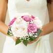 This image captures a romantic, hand-tied bouquet held close to the bride, its serene palette echoing the peaceful lanes and green spaces around Stanmore. Luxurious creamy peonies form the heart of the arrangement, their rounded blooms made of countless soft petals that appear almost weightless. They have the look of flowers freshly gathered from an early morning garden, touched by cool air and soft light. Interwoven with the peonies are roses in complementary tones of dusty pink and soft violet, some with a cooler lilac cast, others edging towards warm rosewood. These roses introduce gentle movement, their spiralling centres drawing the eye deeper into the bouquet's compact, domed silhouette. Delicate wisps of gypsophila emerge between the larger blooms, like a scattering of tiny stars or the faintest hint of bridal veil, bringing a sense of purity and tradition to the design. Shiny dark green leaves cradle the flowers, creating a natural cocoon that sets off the pale petals and gives the bouquet definition when seen against a wedding dress. The stems are likely clasped in a simple ribbon wrap, discreet and elegant. The mood this bouquet conveys is one of quiet sophistication and deep affection, perfectly suited to a ceremony in a Stanmore synagogue, church, or registry office. It suggests fresh, carefully handled blooms, expertly conditioned so they last from morning photos in the garden to evening celebrations. Every element-the soft textures, the pastel hues, the airy baby's breath-works together to mirror the calm, romantic atmosphere couples often seek for their Stanmore wedding day.
