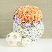 A dense, dome-like cluster of approximately two to three dozen apricot and soft peach roses forms the visual centrepiece here, each flower arranged to show a mix of fully opened blossoms and compact, velvety buds. Threaded among the roses are white Lisianthus blossoms edged in deep purple, their ruffled rims offering a striking, elegant contrast that gives the arrangement a slightly vintage, painterly air. Wisps of Gypsophila (baby's breath) are woven through the bouquet, contributing delicate white specks that lighten the overall mass and lend a timeless softness. The flowers are set into a contemporary rounded white woven vase - an airy lattice of reed or wicker that cradles the blooms like a sculpted nest and adds a tactile, handcrafted texture. Two pale wooden or bamboo decorative spheres sit to the left on the smooth white surface, their open-weave pattern echoing the basket and creating a pleasing rhythm in the composition. The background is a subtle linen-beige wall, softly out of focus so the eye stays with the floral shapes; a soft daylight glow suggests this piece might rest on a Stanmore High Street dining table or brighten a hall near Bentley Priory. Crafted with a local florist's attention to balance and scent, this arrangement, prepared with the care you'd expect from Flowers Stanmore, feels celebratory yet composed - an ideal choice for wedding tables, elegant home decor, or a curated gift for someone who loves quiet luxury.