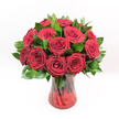 A dozen or more deep red roses stand proudly in the centre of the image, captured against a pure white studio background that makes every petal and leaf seem to glow. Each rose is fully open yet still holds a gentle curl at the heart, with velvety crimson petals layered in tight spirals that suggest romance and devotion. Between the blooms, glossy dark green foliage, likely Ruscus, offers a polished contrast, its pointed leaves framing the bouquet and rounding out the silhouette. The roses are gathered in a translucent ruby-red glass vase with a softly fluted, almost hourglass shape; faint ribbed swirls in the glass catch the light and echo the curves of the petals above. Through the coloured glass, you can see the straight lines of the stems overlapping, grounding the bouquet with a touch of organic structure. Bright, even lighting-like a clear afternoon in a Stanmore sitting room-picks out every nuance of colour and texture, from the soft sheen on the leaves to the satin richness of the petals. The arrangement feels deliberate and composed, as if placed on a dining table in a home just off The Broadway, ready to welcome someone special. It suggests classic Valentine's Day romance, an anniversary surprise, or a heartfelt apology carried through the streets of Stanmore. The image quietly conveys the skill of a professional florist who understands balance, proportion, and how to let red roses speak of love without any need for extra decoration or distraction.
