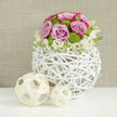 A meticulously crafted floral centerpiece stands on a snow-white, lightly reflective tabletop, the kind you might find in a contemporary Stanmore home set for an intimate wedding celebration. The central focus is a spherical vase created from white-painted natural branches, woven together to form a delicate, open lattice that feels simultaneously rustic and modern. Cradled inside is a lush, dome-shaped arrangement of flowers, with pastel pink roses taking pride of place. These roses are fully open, their velvety petals spiralling outward, some delicately tipped with a richer pink that adds a romantic nuance to the overall palette. Threaded throughout are small lime-green button chrysanthemums, their compact, rounded blooms creating a lively contrast in both colour and texture, like tiny bursts of fresh spring grass among the blossoms. Encircling the lower edge of the bouquet, a fringe of small white flowers with subtle yellow centres tumbles gently over the woven rim, softening the structure and creating a graceful transition from flowers to vase. To the left side of the arrangement, two decorative wooden orbs made from pale, flat strips of wood rest on the table: one is larger, one smaller, and their open, basket-like design mirrors the texture of the vase, adding another note of organic charm. Behind, a softly blurred, warm neutral wall in beige or taupe provides a calm, textured backdrop that lets the colours and shapes come forward clearly. The entire composition radiates considered elegance and quiet joy, ideal for wedding centerpieces at local venues near Stanmore station, as well as anniversary dinners or special family gatherings in the area.
