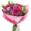 A radiant, hand-tied bouquet sits upright in a clear cylindrical glass vase, its bundled stems visible beneath water and a simple loop of rustic twine. At the visual heart, several large gerbera daisies command attention in a vivid fuchsia pink, their densely layered petals forming circular spokes around dark, almost black centers that give the blooms a striking, graphic contrast. Nestled among them are multiple soft pink roses in a range of openings - from tight, elegant buds to lush, fully opened blossoms with gently unfurling layers - lending a romantic, tender counterpoint to the gerberas' boldness. Lush greenery, a mix of broad, smooth leaves and slender stems, creates a fresh, verdant backdrop that adds depth and a living, natural frame. Sprays of deep purple-blue filler flowers are scattered through the composition, their fine texture drawing the eye inward and adding subtle shadow and richness to the palette. The bouquet is wrapped in crinkled, sheer pink cellophane or tissue that graduates from an almost translucent pastel halo at the top to a more saturated, brighter pink toward the base, catching the light with soft gleams and creating a voluminous, celebratory skirt. A small white rectangular tag hangs from the twine at the vase neck, printed with a stylised pink-and-green rose emblem that suggests a florist's signature. The overall effect is fresh, cheerful and uplifting - a composition that reads as both carefully designed and effortlessly joyful, ideal for birthdays, anniversaries or to brighten a room with the feeling of a spring morning in Stanmore.