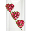 Three lush, rose-filled hearts cascade diagonally across a smooth white background in this expressive funeral tribute, each heart brimming with roses in deep red, bright fuchsia, and gentle pink that together create a vivid yet tender palette. The roses are tightly massed, so that the viewer sees almost nothing but petal upon petal, shaped into perfect hearts that feel both strong and delicate at the same time. Some flowers curl inward protectively, while others open generously toward the light, building a sense of depth and movement across the surface of each heart. Tracing their outlines, slender ivy vines sweep in gentle curves, adding a flowing green frame that continues downward to join one heart to the next. The ivy leaves, small and glossy, introduce a calming natural contrast to the passion of the red roses, like a whisper of a garden in Stanmore on a quiet afternoon near Canons Park. The clean, white background recalls a modern memorial setting, perhaps a service space close to Stanmore's leafy residential streets, where families gather to say farewell with dignity and love. Soft lighting illuminates the arrangement evenly, bringing out the richness of the roses' colours and the sheen of the foliage without harshness, maintaining a peaceful tone. This triple-heart design would be especially meaningful as a symbol of close family ties-a tribute from children, grandchildren, or a devoted partner-delivered to local Stanmore venues or chapels. Each heart reads as a distinct message of love, yet together they form a single, flowing statement of remembrance, expressing how deeply the departed is held in the hearts of those who remain.