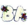 A visually striking and deeply respectful floral tribute forms the capital letters BA, set side-by-side on a low, dark green rectangular base against a clean white background. Each letter is built from hundreds of small, pristine white chrysanthemum florets pressed close together to create a soft, pillowy surface that reads as both delicate and durable. The edges of the B and A are neatly trimmed and framed with a ruffled royal blue satin ribbon, which catches light and gives each letter a crisp, defined silhouette while emphasising the contrast between the pure white face and the vibrant border.
Accents are placed to balance the composition: a lush cluster of velvety deep red roses adorns the upper left curve of the B, and a matching cluster sits at the lower right of the A. Interspersed among the roses are vivid, spike-like blue delphinium-style blooms and small sprays of lighter green trailing foliage that soften the geometry and add a whisper of movement. Darker green leaves tuck behind the blossoms to provide depth and a natural frame. Under soft studio light the white chrysanthemums glow quietly and the roses release a faint, classic perfume, lending the piece a calm, contemplative mood. Crafted with the measured restraint used for memorial work, this pair of initial letters is tailored for a funeral or commemorative service, suitable for display at local chapels, community halls, or quiet corners near Stanmore Common, where families seek a personalised and dignified way to honour a loved one.
