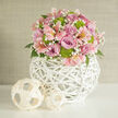This image captures a carefully crafted floral centerpiece that would look perfectly at home on a wedding table in Stanmore or a stylish dining room near Belmont Circle. The bouquet is shaped into a low, rounded dome, every flower nestled closely to create a harmonious, seamless effect. At its core, full-headed roses in shades of lilac and warm pink unfurl their velvety petals, each bloom overlapping the next in a rich, romantic cluster. Threaded among them, pale pink alstroemeria open with long, graceful petals touched by fine freckles and gentle stripes, adding both subtle pattern and movement. Bright lime-green button chrysanthemums appear like little bursts of freshness, their tidy, spherical forms punctuating the softer colours and lending the arrangement a clean, modern edge. Wisps of white baby's breath weave through the design, their tiny, airy blossoms acting like a delicate veil that softens the overall look and allows light to flicker between the larger flowers. Dark green foliage is mostly hidden beneath, but occasional leaves peek through, framing the pastel tones with a natural, grounded base. All of this floral detail is gathered into a striking white woven vase, its spherical shape and open, rattan-like lattice turning the container into a sculptural accent rather than a mere support. The vase sits on a spotless white surface, where its curves and the lower petals of the bouquet catch and reflect the ambient light. Beside it, two smaller decorative spheres in a paler, natural wood weave mirror the pattern of the vase, tying the whole arrangement together visually. The background is a soft, neutral beige wall, unobtrusive and calm, ensuring the bouquet remains the clear focal point. The overall impression is one of gentle celebration, freshness, and thoughtful design, ideal for marking special moments and same-day floral delivery throughout Stanmore.