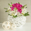 A compact yet luxurious floral arrangement in a white wicker basket appears as though it has been styled for a refined Stanmore wedding table. The design is low and rounded, its silhouette smooth and perfectly proportioned. At its heart, lustrous fuchsia blooms cluster closely together, their densely packed petals curving inwards and outwards to form ruffled, almost cushion-like shapes. Their vivid, jewel-toned colour provides a striking contrast to the ring of ivory alstroemeria surrounding them. The alstroemeria blossoms are soft and light, with slim trumpet-shaped petals that flare open gently, marked with fine touches of pink and subtle speckling that add delicate character to each flower. Interspersed among the blossoms, small buds and fresh green leaves emerge, punctuating the arrangement with hints of growth and the promise of new chapters, much like couples beginning married life in Stanmore's leafy streets. 

The flowers are nestled in a round basket crafted from bright white wicker, its interwoven strands forming an intricate lattice with a modern, sculptural feel. The weave is open enough to let light pass through, casting tiny shadows on the surface beneath and emphasizing the hand-crafted nature of the piece. Two small, cream-coloured orbs made from similar woven material lie close by, echoing the texture of the basket and giving the overall composition a gentle, curated look. The lighting is soft and even, reminiscent of natural daylight streaming through large windows in a Stanmore home. The mood is polished yet warm, ideal for wedding centrepieces at local venues, civil ceremonies near Stanmore station, or elegant thank-you flowers delivered to family after the celebration. The imagined scent is fresh and green with a delicate floral sweetness, never overpowering, simply enhancing the sense of quiet joy.