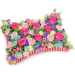 A pillowy, rectangular floral tribute shown from a gentle overhead oblique angle against a crisp white backdrop, its surface densely packed like a soft woven cushion of blooms. The dominant hot-pink roses read as velvety focal points, some petals tightly furled and others unfurling into plush spirals; around them pale shell-pink spray carnations provide a ruffled, tactile softness. Scattered through the composition are deep purple lisianthus with their bell-like faces and cheerful lemon-yellow ranunculus that give the piece lively highlights, while tiny pale-pink filler blooms knit the groups of flowers together. Architectural contrast is supplied by a few spiky, silver-blue Eryngium thistles, whose bristling texture and steelier tone cool the palette and add a wild, sculptural line against the rounded roses. Rounded, silvery-green eucalyptus leaves and darker green foliage form a verdant underlayer, their matte, cool surfaces giving depth and a fresh, aromatic lift that I picture smelling faintly of citrus and camphor. The whole cushion rests within a pleated deep-red satin border, its ruffled trim neatly defining the pillow silhouette and lending quiet dignity to the arrangement. This is the kind of sympathy pillow one might place near a service at a Stanmore chapel or at a graveside in Stanmore Park - a composition that celebrates memory with colour rather than sombre restraint. The studio lighting accentuates petals' subtle sheen and cast soft, minimal shadows, emphasizing the careful density and balance of the design. As a local florist might note, the piece reads as both comforting and celebratory: an intimate, crafted expression of remembrance familiar to families around Stanmore Broadway and the tube station.