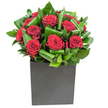 In this elegantly modern composition, a cluster of velvety crimson roses forms the immediate focal point, each bloom unfurling in plush, spiral layers that read as both intimate and dramatic. The roses are nestled into broad, glossy green leaves that have been deliberately curled and overlapped to create a rhythmic sculptural frame - the foliage's waxy sheen catches the light and gives the display lively movement and depth. The arrangement sits low and confident in a matte black, square box vase whose clean geometry grounds the composition and provides a contemporary counterpoint to the softness above. Lighting is bright and clear, the kind of steady studio glow that makes the crimson tones glow like velvet and throws gentle highlights along the ridges of each petal and the glossy margins of the leaves. If you lean in, the scent is subtle: classic rose sweetness lifted by a cool, green freshness that hints at clipped garden foliage rather than heavy perfume. Tactile contrasts are obvious even from a distance - the silken petals against the firm, almost tropical texture of the leaves - and the overall balance reads as carefully considered, meant to be both seen and felt. In a local context, this is the kind of bouquet that would add quiet drama to a Stanmore supper table, make a romantic milestone feel decisive, or arrive as a thoughtful expression of thanks after an evening at Canons Park. Crafted with intent and modern restraint, the arrangement speaks of affection, composed with the kind of care you'd expect from an experienced florist serving Stanmore and nearby Harrow.