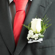 An ivory rose, cultivated to a soft, velvety perfection, forms the focal point of this boutonnière, set against the smart, matte charcoal of a jacket and the subtle pattern of a checked shirt beneath. Tiny clusters of baby's breath surround the rose like a delicate halo, their pinpoint blooms adding air and innocence to the arrangement while tempering the rose's quiet strength. Narrow, dark-green leaves and slender curled blades arch gracefully outward, their glossy surfaces providing structural contrast and a cooling visual frame. The composition is finished with a slim white satin ribbon tied in a restrained bow at the base, introducing a hint of ceremonial shine that echoes the silk of a red tie when paired for a groom or honored guest. In soft daylight the textures resolve into a calm study of tone - creams, deep greens, and charcoal - and the scent is subtle: rose-sweet with a green, leafy underpinning that feels freshly cut and dew-sifted. As a local florist familiar with Stanmore's weddings and civic events, we design this piece to speak to both tradition and contemporary minimalism, confident that it will sit comfortably beside a tailored suit whether the ceremony is near the High Street shops or along the leafy approaches to Canons Park. Thoughtfully proportioned and cool to the touch, this boutonnière reads as understated elegance: an intimate accent crafted to be felt as much as seen, a small but meaningful emblem of presence and celebration.